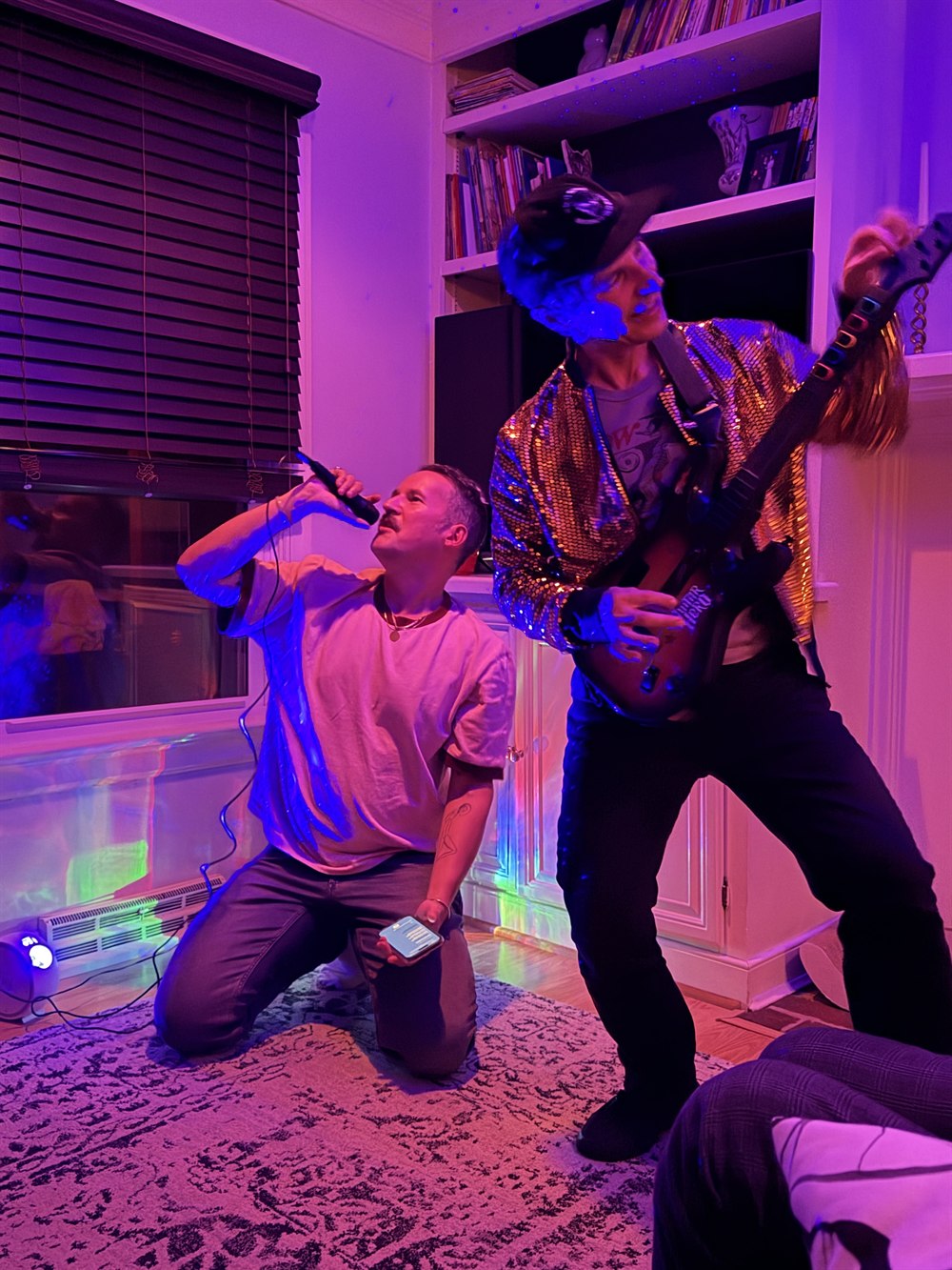 Two friends in a living room lit purple and blue by party lights. One kneels on the rug singing into a microphone with his head back; beside him the host, in a gold sequin jacket and a black cap, plays a plastic guitar controller with one arm thrown up. Bookshelves and LED strips glow behind them.