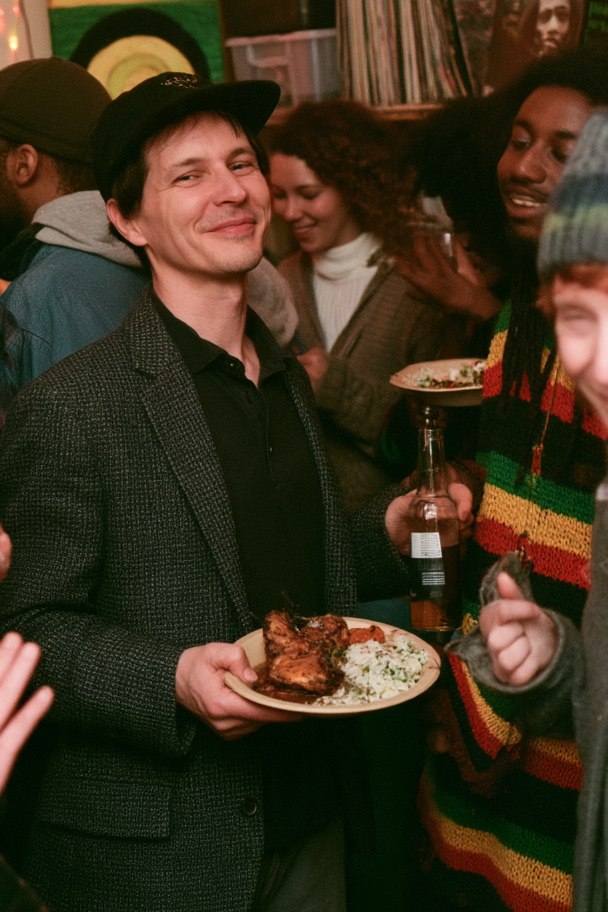 The host near the end of the night in a flat cap and tweed jacket, holding a plate of jerk chicken and rice, smiling. Red and green light, dub reggae posters and a speaker cabinet behind him, guests still talking around him.