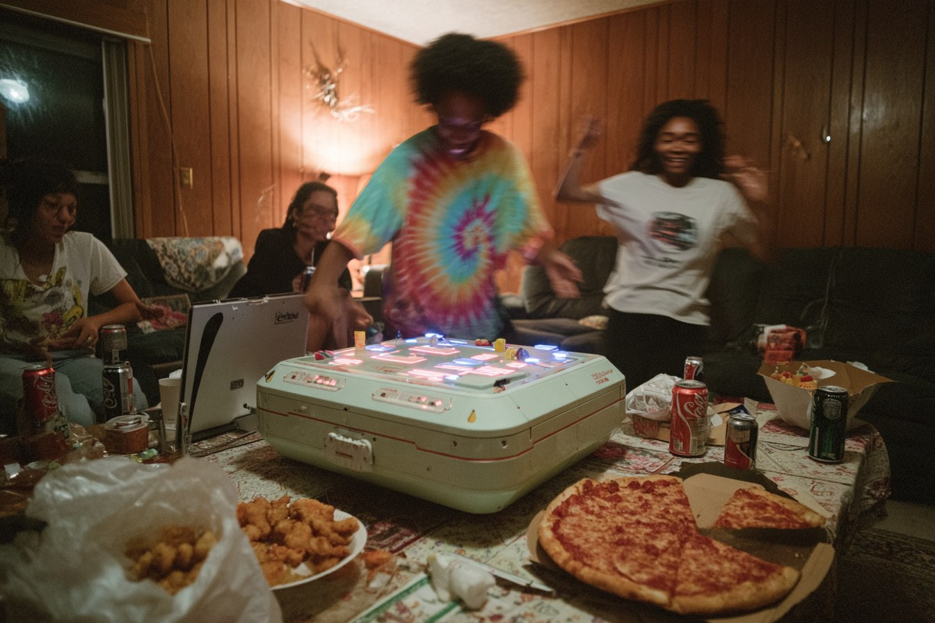 A wood-panelled basement rec room. A large pale drum machine the size of a suitcase sits on the coffee table, its pads glowing pink and blue. A woman in a tie-dye shirt leans over it mid-motion while another dances behind her. A whole pizza, a plate of tater tots and scattered soda cans surround it.